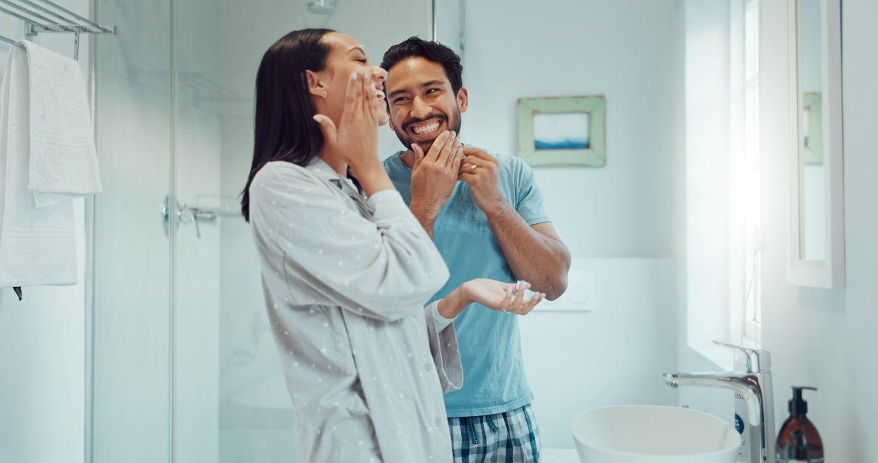 Laechelndes Paar im Badezimmer mit Dusche und schlichter Duschrueckwand im Hintergrund